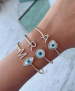 Load image into Gallery viewer, A stack of bangles on a wrist, featuring different designs including a customizable initial and an evil eye charm in gold color.