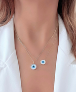 Load image into Gallery viewer, A person wearing a white shirt showcasing two necklaces, one with a round mother of pearl charm and a smaller evil eye charm, both with gold trim, against their neck.