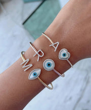 A stack of bangles on a wrist, featuring different designs including a customizable initial and an evil eye charm in gold color.